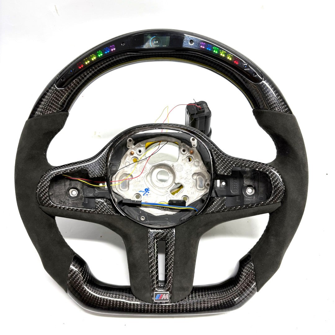 CJ Mod Garage BMW G-chassis LED carbon fiber steering wheel on white background featuring integrated RGB LED shift indicators, digital top display module, gloss carbon fiber shell, Alcantara side grips, flat-bottom racing profile, exposed OEM mounting structure, compatible with BMW G02 G05 G06 G09 G11 G12 G15 G20 G22 G28 G29 G30 G38 G80 G82 F40 F90