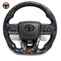 Toyota LC300 steering wheel featuring blue forged carbon material black perforated leather side grips integrated buttons and GR sport flat bottom styling by CJ Mod Garage