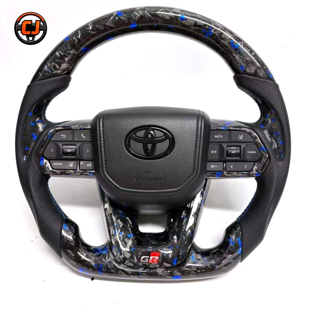 Toyota LC300 steering wheel featuring blue forged carbon material black perforated leather side grips integrated buttons and GR sport flat bottom styling by CJ Mod Garage