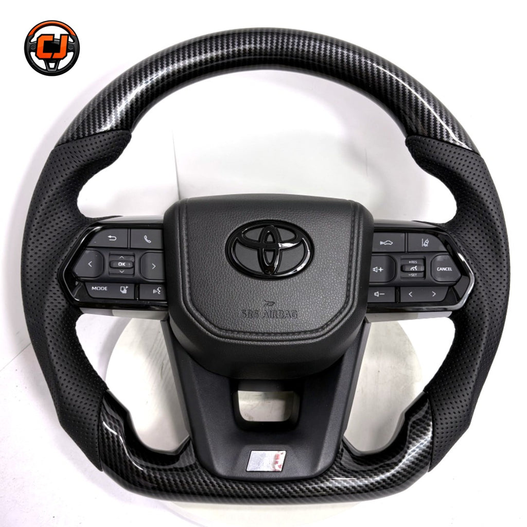 Toyota LC300 premium steering wheel with hydro-dipped carbon fiber pattern, perforated leather side sections, factory airbag cover and integrated controls by CJ Mod Garage