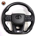 Toyota LC300 premium steering wheel with hydro-dipped carbon fiber pattern, perforated leather side sections, factory airbag cover and integrated controls by CJ Mod Garage