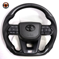 Toyota LC300 premium steering wheel with hydro-dipped carbon fiber pattern, perforated leather side sections, factory airbag cover and integrated controls by CJ Mod Garage