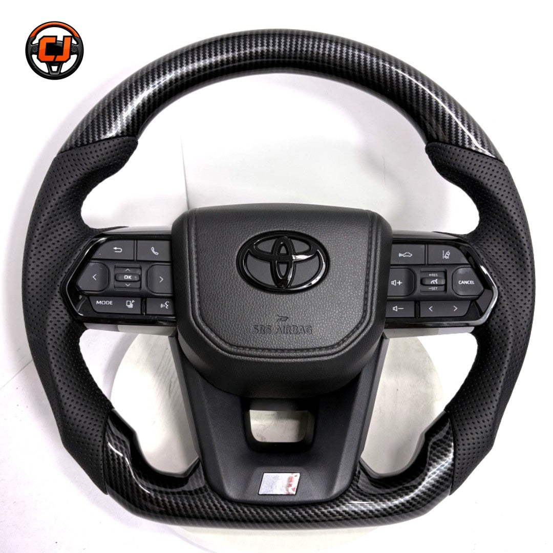 Toyota LC300 premium steering wheel with hydro-dipped carbon fiber pattern, perforated leather side sections, factory airbag cover and integrated controls by CJ Mod Garage