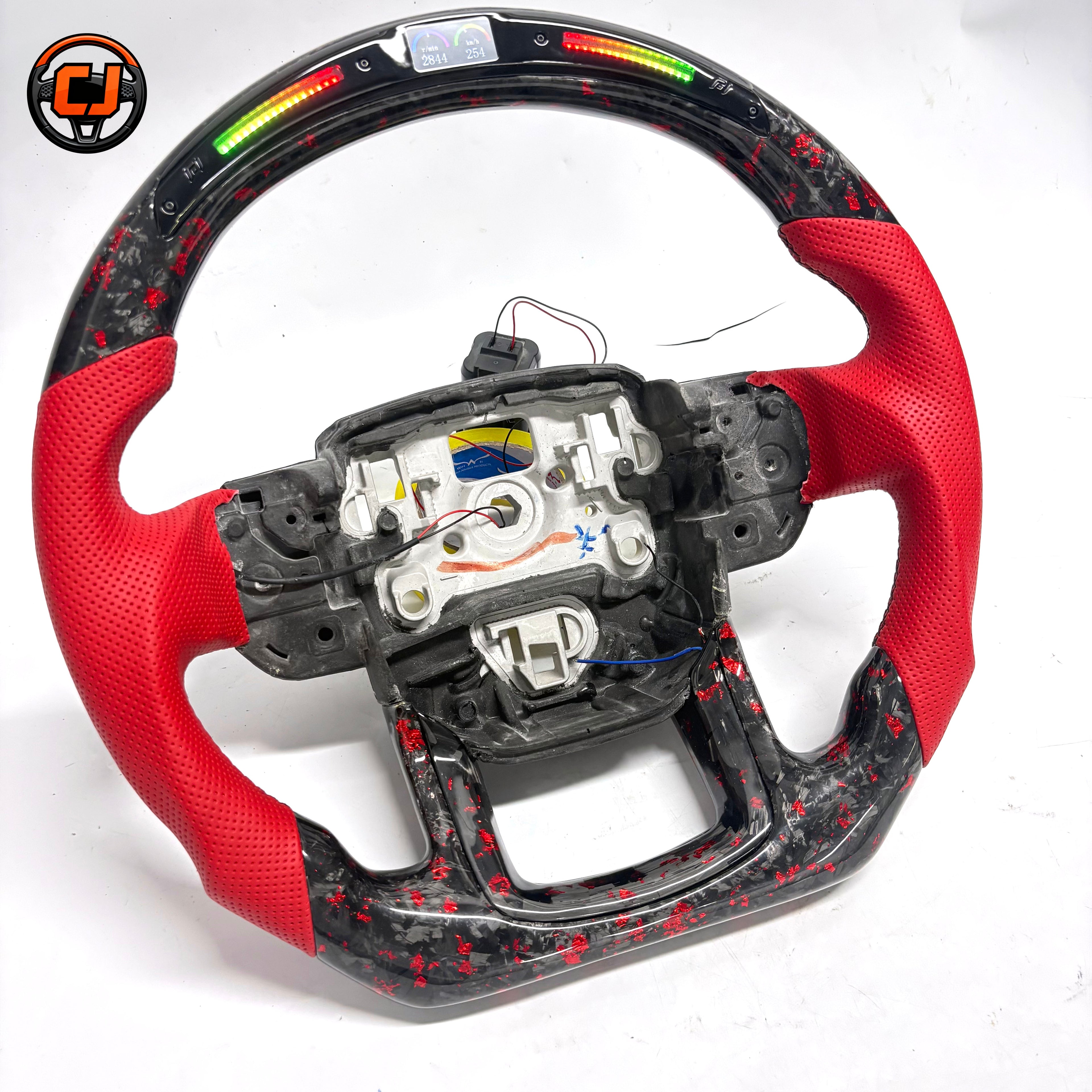 Studio product photo of custom Land Rover Range Rover carbon fiber steering wheel with red perforated leather grips, LED shift lights, top digital display and exposed airbag mounting structure