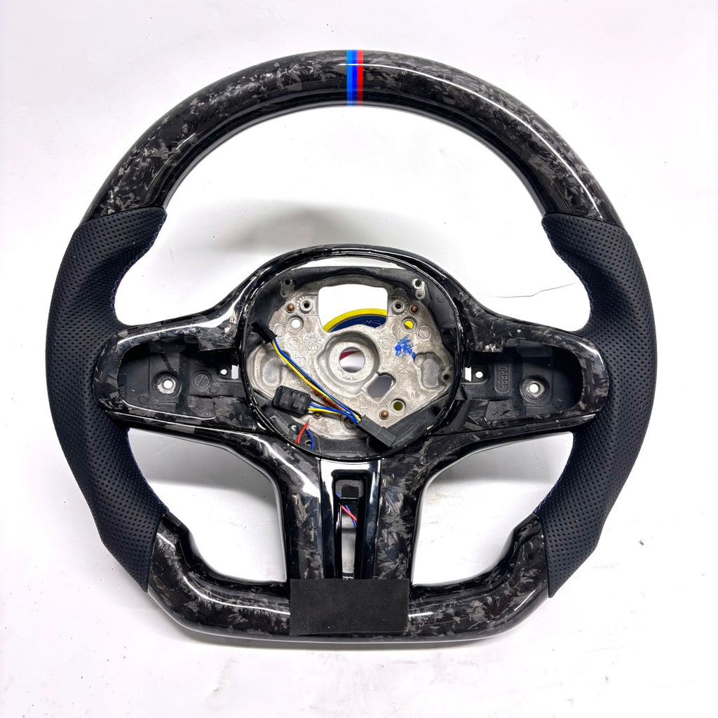CJ Mod Garage BMW G-chassis forged carbon fiber steering wheel on white background featuring gloss forged carbon construction, perforated black leather side grips, flat-bottom performance shape, M tricolor 12 o’clock stripe, exposed mounting structure for OEM buttons and airbag components, compatible with BMW G02 G05 G06 G09 G11 G12 G15 G20 G22 G28 G29 G30 G38 G80 G82 F40 F90