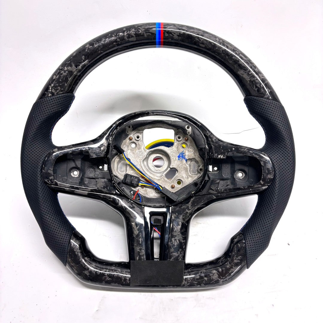 CJ Mod Garage BMW G-chassis forged carbon fiber steering wheel on white background featuring gloss forged carbon construction, perforated black leather side grips, flat-bottom performance shape, M tricolor 12 o’clock stripe, exposed mounting structure for OEM buttons and airbag components, compatible with BMW G02 G05 G06 G09 G11 G12 G15 G20 G22 G28 G29 G30 G38 G80 G82 F40 F90