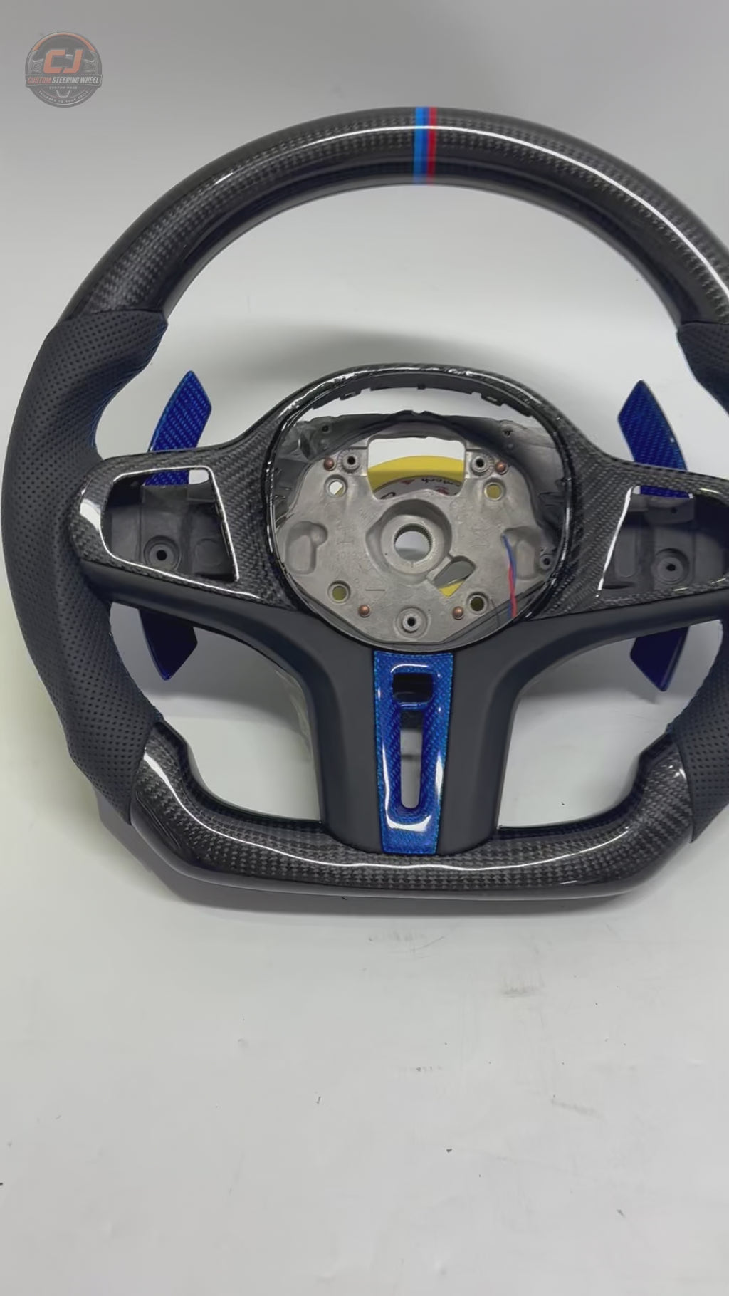 White background product showcase video of CJ Mod Garage BMW custom carbon fiber steering wheel featuring gloss forged carbon fiber construction, black perforated leather grips, blue forged carbon accents, M tricolor 12 o’clock stripe, flat-bottom motorsport design, OEM-style fitment, and compatibility with BMW G02 G05 G06 G09 G11 G12 G15 G20 G22 G28 G29 G30 G38 G80 G82 F40 F90