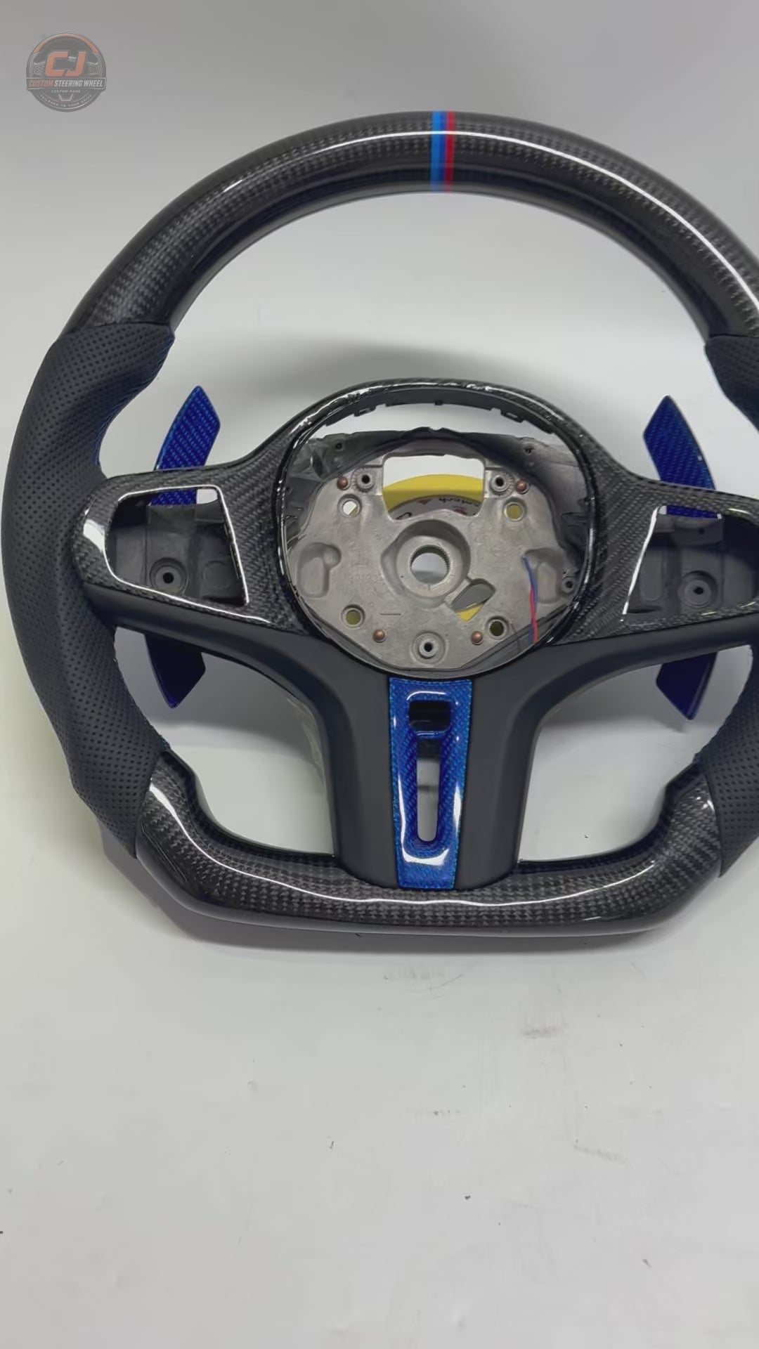 White background product showcase video of CJ Mod Garage BMW custom carbon fiber steering wheel featuring gloss forged carbon fiber construction, black perforated leather grips, blue forged carbon accents, M tricolor 12 o’clock stripe, flat-bottom motorsport design, OEM-style fitment, and compatibility with BMW G02 G05 G06 G09 G11 G12 G15 G20 G22 G28 G29 G30 G38 G80 G82 F40 F90