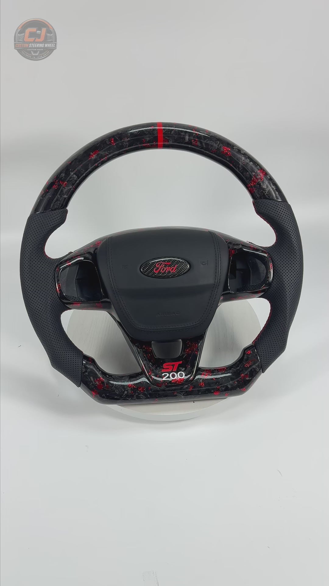 Studio product video showcasing a Ford Focus ST200 forged carbon fiber steering wheel with red metallic flakes, perforated leather grips, red alignment stripe, flat-bottom design and custom detailing by CJ Mod Garage