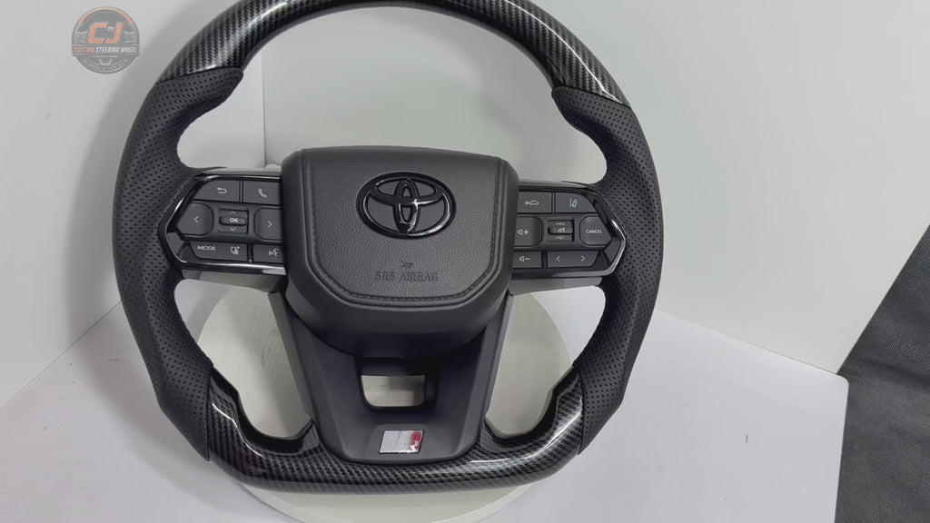 White background showcase video of Toyota Land Cruiser LC300 hydro-dipped carbon style steering wheel with perforated leather grips, OEM control buttons and factory-style fitment by CJ Mod Garage