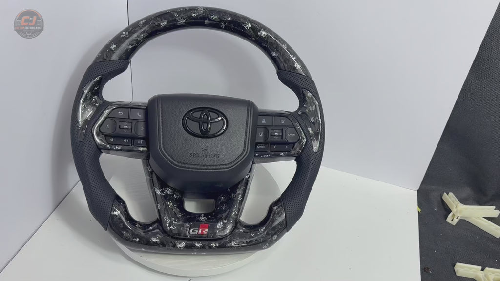360-degree product video of Toyota LC300 forged carbon steering wheel with white flake finish, perforated leather grips, OEM button layout and plug-and-play fitment by CJ Mod Garage