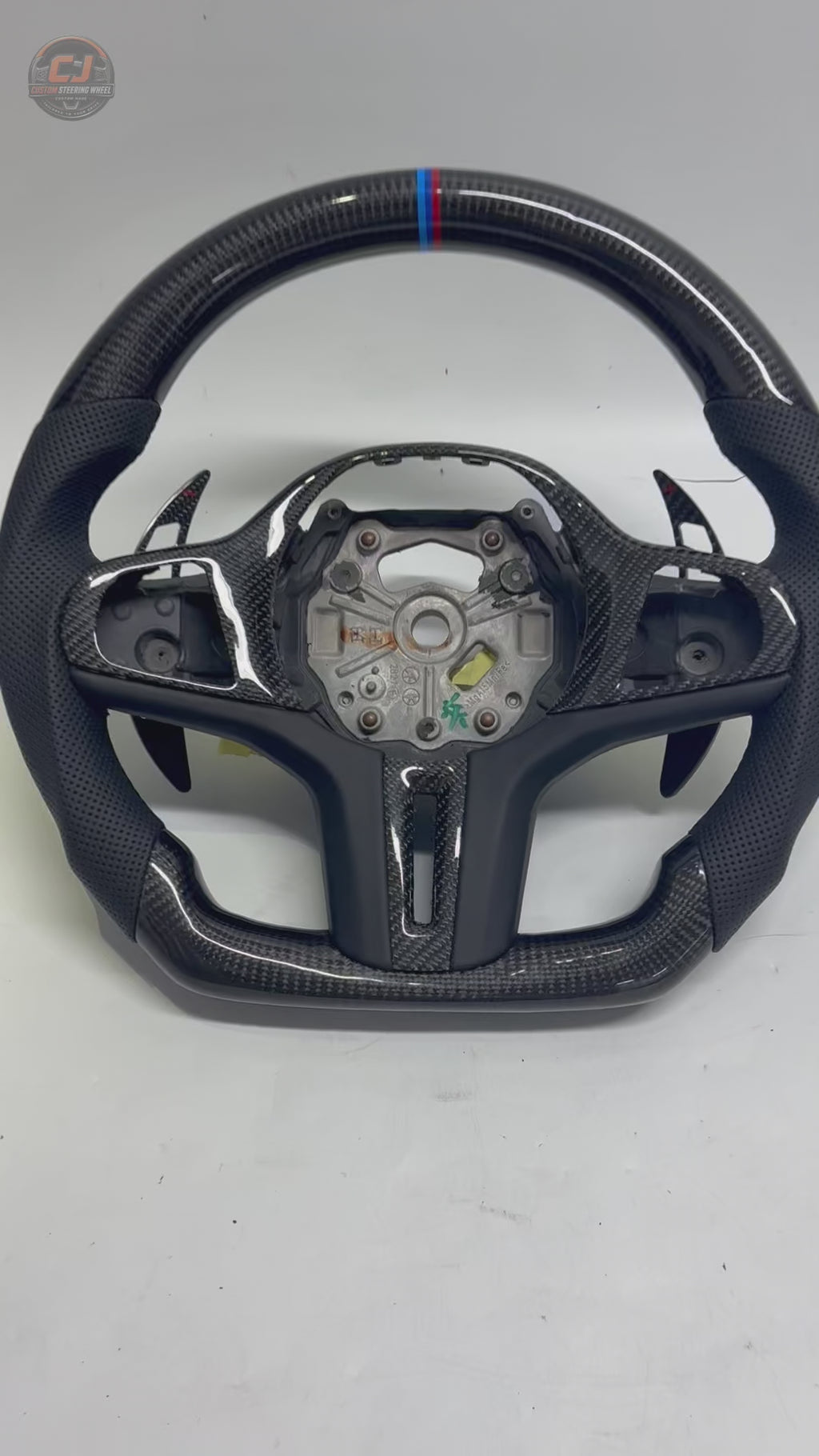 White background showcase video of CJ Mod Garage BMW custom carbon fiber steering wheel featuring gloss forged carbon fiber construction, black perforated leather grips, M tricolor 12 o’clock stripe, flat-bottom performance design, OEM-inspired styling, and compatibility with BMW G02 G05 G06 G09 G11 G12 G15 G20 G22 G28 G29 G30 G38 G80 G82 F40 F90