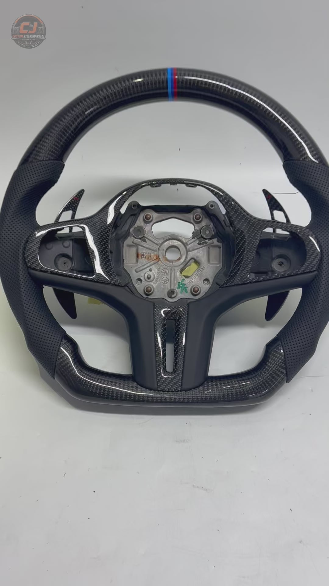White background showcase video of CJ Mod Garage BMW custom carbon fiber steering wheel featuring gloss forged carbon fiber construction, black perforated leather grips, M tricolor 12 o’clock stripe, flat-bottom performance design, OEM-inspired styling, and compatibility with BMW G02 G05 G06 G09 G11 G12 G15 G20 G22 G28 G29 G30 G38 G80 G82 F40 F90