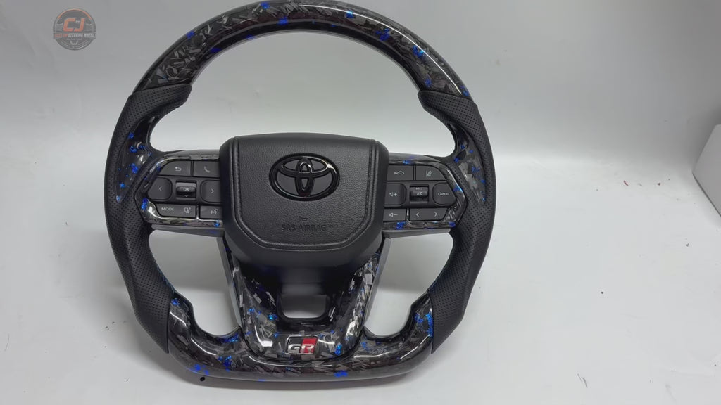 White background rotating video of Toyota LC300 steering wheel showcasing blue forged carbon material perforated leather grips GR sport design OEM controls and custom craftsmanship by CJ Mod Garage