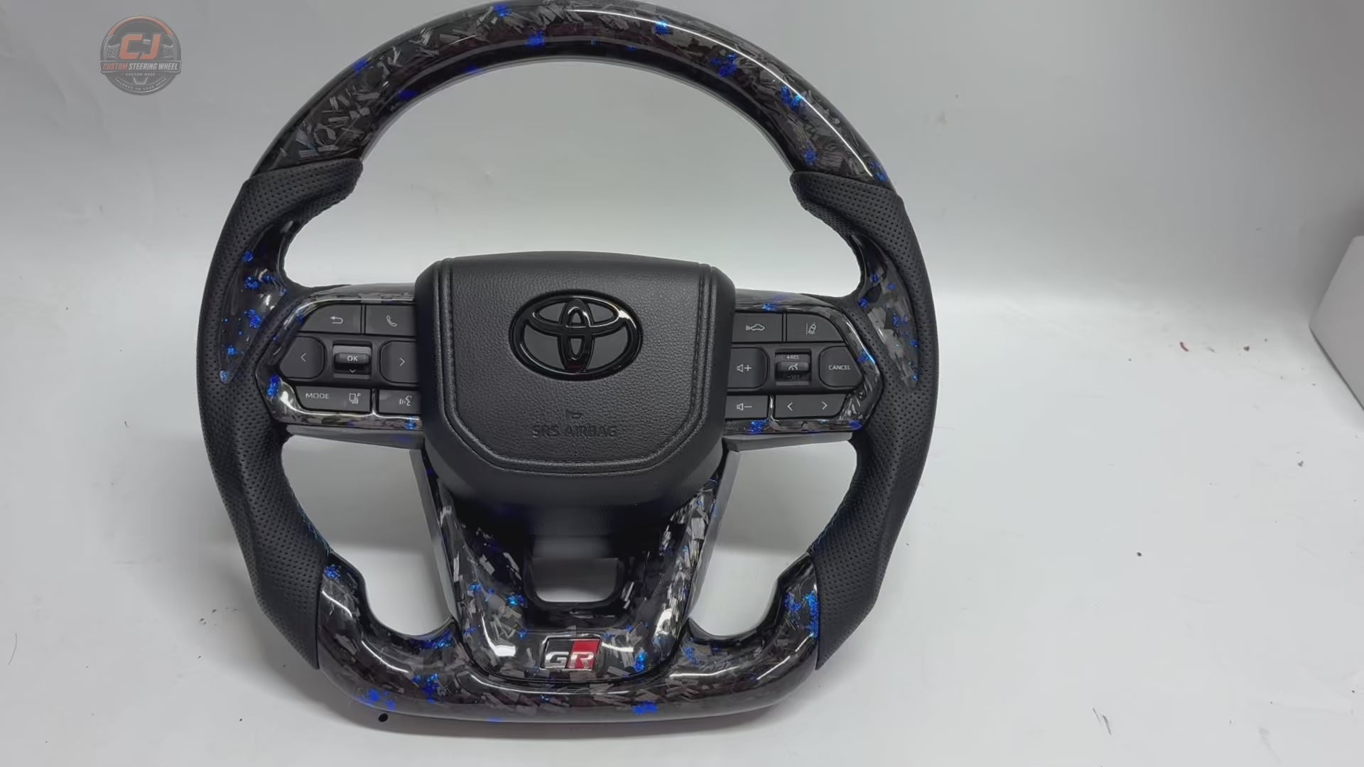 White background rotating video of Toyota LC300 steering wheel showcasing blue forged carbon material perforated leather grips GR sport design OEM controls and custom craftsmanship by CJ Mod Garage