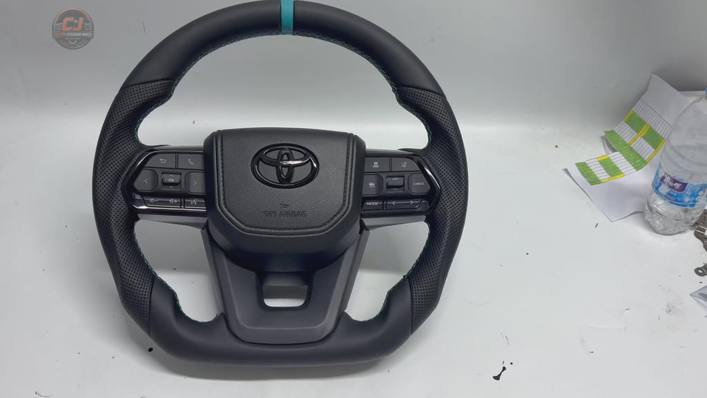 White background product video showcasing Toyota LC300 custom leather steering wheel with perforated leather grips, blue center stripe, OEM multifunction buttons and precision craftsmanship by CJ Mod Garage