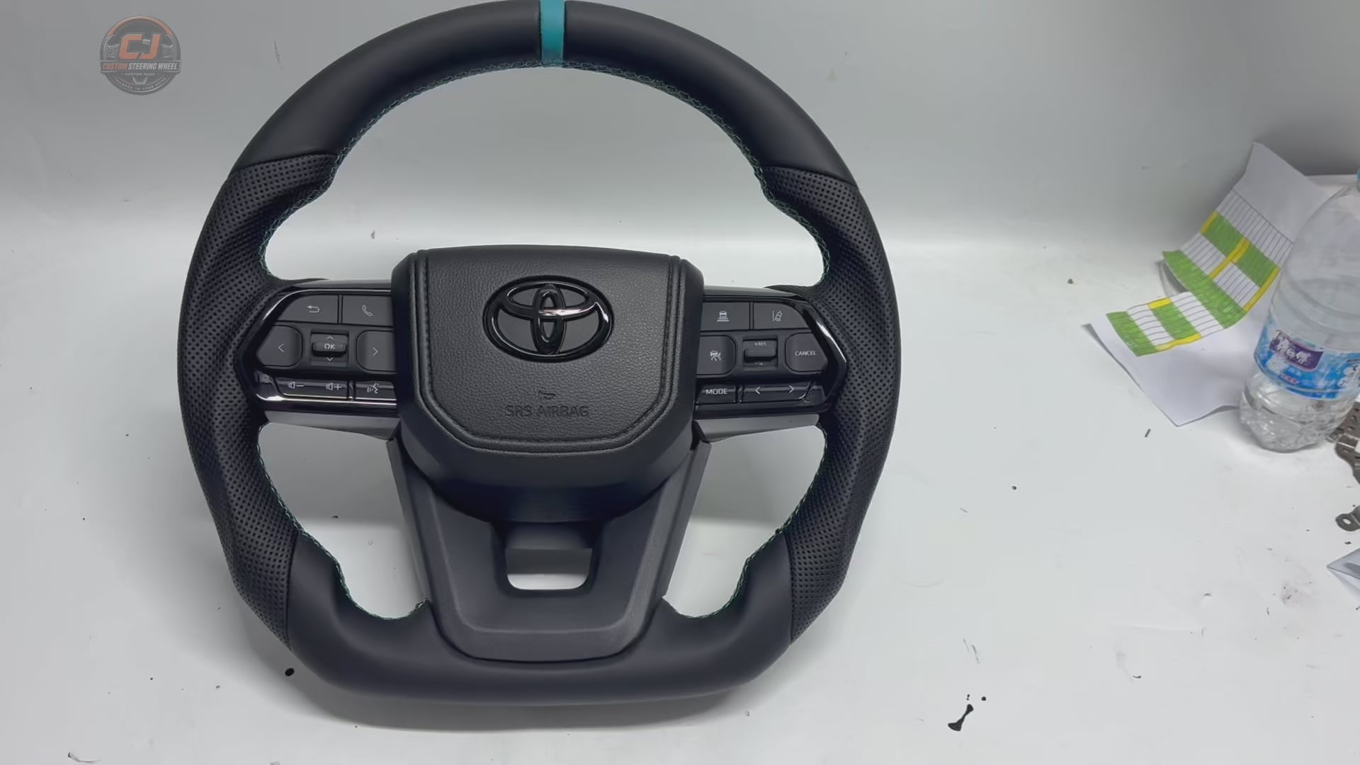 White background product video showcasing Toyota LC300 custom leather steering wheel with perforated leather grips, blue center stripe, OEM multifunction buttons and precision craftsmanship by CJ Mod Garage