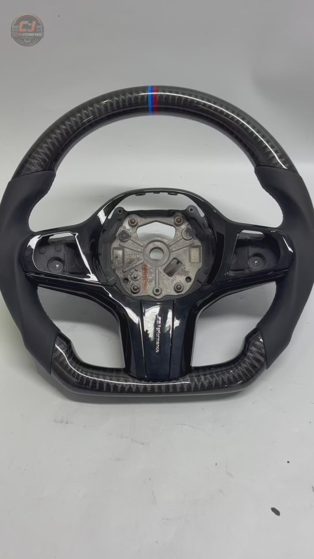 White background product showcase video of CJ Mod Garage BMW custom carbon fiber steering wheel with gloss forged carbon fiber finish, black leather sport grips, gloss black center trim, M tricolor center marker, flat-bottom performance design, and OEM-style fitment for BMW G02 G05 G06 G09 G11 G12 G15 G20 G22 G28 G29 G30 G38 G80 G82 F40 F90